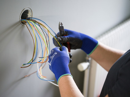 Person med blå handskar arbetar med elkablar i en väggöppning, håller i en tång.