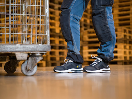Person i blå arbetsbyxor och svarta säkerhetsskor står på lagergolv nära bur på hjul.