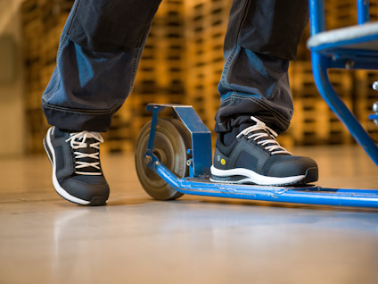 Person med svarta skyddsskor står på en sparkcykel i lagerliknande miljö.