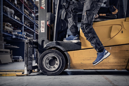 En person i arbetskläder och sneakers sitter på en gul gaffeltruck i ett lager.