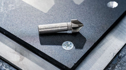 Metallborr ligger på en svart plåt med ett borrat hål bredvid.