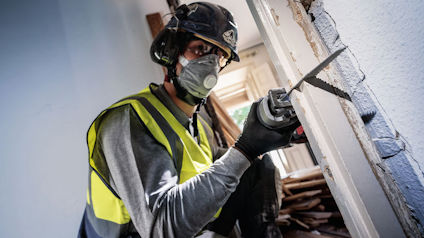 Person i skyddskläder och hjälm använder en eldriven såg för att skära i en dörrkarm.