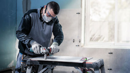 Person arbetar med elverktyg på arbetsbänk inomhus, bär skyddsglasögon och andningsskydd.