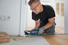 Man in protective eyewear using an electric saw to cut flooring near doorway, with wood panels stacked nearby.