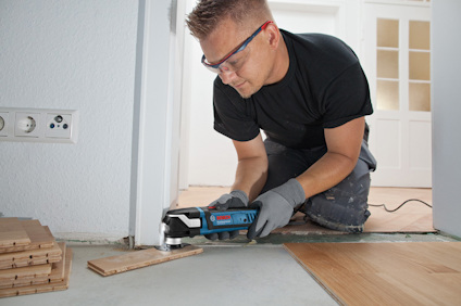 Man in protective eyewear using an electric saw to cut flooring near doorway, with wood panels stacked nearby.