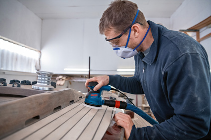Person använder en slipmaskin på en träyta, iförd skyddsglasögon och ansiktsmask i en verkstad.