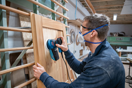 Person slipar träram med en elektrisk slipmaskin. Bär skyddsglasögon och andningsmask.
