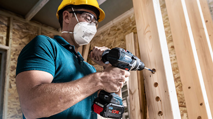 Person med skyddshjälm och mask borrar hål i träbalkar inomhus.