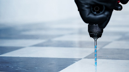 Hand med svart handske borrar i ett schackrutat golv med ett blått borr.