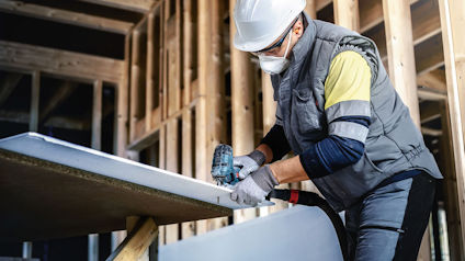 Byggarbetare med hjälm och skyddsglasögon sågar gipsskiva inomhus.