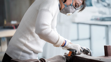 Person i vit tröja och skyddsmask slipar trä med verktyg i en verkstad.