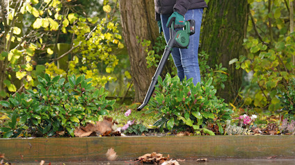 Person använder en grön lövblåsare i en trädgård nära buskar och träd.
