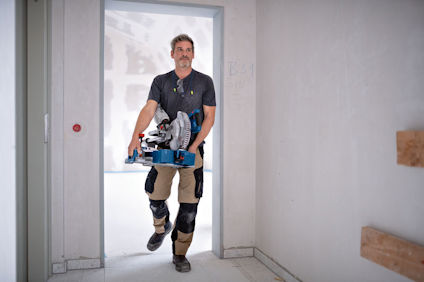 En person bär en bordsåg genom en dörröppning i en byggmiljö.