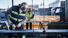Byggarbetare slipar metallstång utomhus, bär skyddshjälm och reflexväst. En annan arbetare syns i bakgrunden.