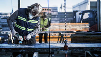 Byggarbetare slipar metallstång utomhus, bär skyddshjälm och reflexväst. En annan arbetare syns i bakgrunden.