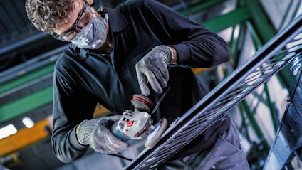 Person med skyddsglasögon och handskar använder en vinkelslip på metallstag i industriell miljö.