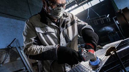 Person i skyddsmask och handskar använder en vinkelslipmaskin i industrimiljö.
