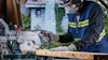 Person som använder en Bosch-kapsåg, bär skyddsmask och handskar, sågar en träbit i utomhusmiljö.