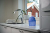 Blå liten behållare på köksbänk nära diskho, staplade handdukar och fönster med utsikt mot trähus.