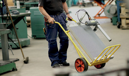 Person med säckkärra bär blålackerad komponent och verktyg med ormliknande rör i industriell verkstad.