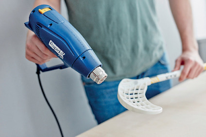 En person håller en blå värmepistol och en vit plastbit med rutmönster.